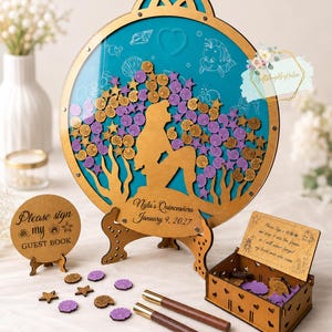 Può includere: Un libro degli ospiti in legno a tema sirena, con sfondo turchese e dettagli dorati. Il design include una silhouette di sirena, decorazioni floreali e a stella e il testo "Nyla's Quinceañera January 9, 2027". Include un cartello, penne e una scatola.