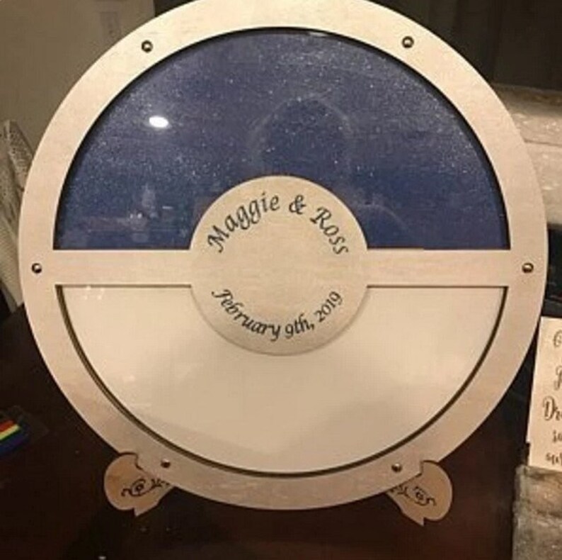 K&ouml;nnte beinhalten: Eine runde Holzkiste mit einem blauen und wei&szlig;en geteilten Innenraum. Die Kiste hat einen durchsichtigen Glasdeckel und einen kleinen Holzst&auml;nder. Die Kiste ist mit dem Text "Maggie & Ross February 9th, 2019" graviert.