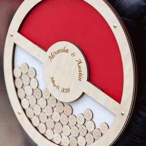 K&ouml;nnte beinhalten: Eine h&ouml;lzerne Hochzeitskartenbox mit rotem und wei&szlig;em Design. Die Box ist in Form eines Pok&eacute;balls gestaltet und tr&auml;gt die Namen "Miranda & Austin" sowie das Datum "May 6, 2018" eingraviert.