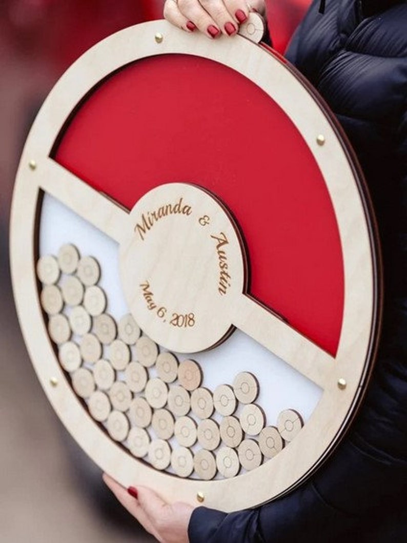 K&ouml;nnte beinhalten: Eine h&ouml;lzerne Hochzeitskartenbox mit rotem und wei&szlig;em Design. Die Box ist in Form eines Pok&eacute;balls gestaltet und tr&auml;gt die Namen "Miranda & Austin" sowie das Datum "May 6, 2018" eingraviert.