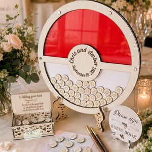 Puede incluir: Un libro de visitas de boda de madera con un dise&ntilde;o circular rojo y blanco. El libro de visitas tiene un marco circular con un fondo rojo y un espacio para que los invitados firmen fichas de madera. Los nombres "Chris y Amber" y la fecha "15 de septiembre de 2022" est&aacute;n grabados.