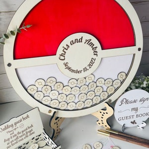 Puede incluir: Un libro de visitas de boda de madera con forma de c&iacute;rculo rojo y blanco con los nombres "Chris and Amber" y la fecha "September 15, 2022". Incluye una caja de fichas de madera para que los invitados firmen y dejen caer en el libro. Un letrero dice "Please sign my GUEST BOOK".