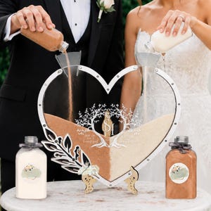 May include: A heart-shaped sand ceremony set with a clear acrylic frame and wooden stand. The set includes two bottles of sand being poured into the frame, and two additional bottles of sand. The frame has a tree and initials design.