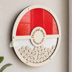Puede incluir: Un libro de visitas alternativo redondo de madera con un dise&ntilde;o rojo y blanco. La secci&oacute;n superior es roja y la secci&oacute;n inferior es blanca. El centro tiene los nombres "Chris y Amber" y la fecha "15 de septiembre de 2022". Peque&ntilde;os c&iacute;rculos de madera debajo.