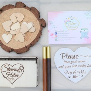 K&ouml;nnte beinhalten: Eine wei&szlig;e Holzkiste mit einem herzf&ouml;rmigen Design und dem Text "Steven & Helen September 09, 2023". Die Kiste ist umgeben von Holzh Herzen, zwei Holzstifte und einem wei&szlig;en herzf&ouml;rmigen Schild mit der Aufschrift "Bitte hinterlassen Sie Ihren Namen und Ihre besten W&uuml;nsche f&uuml;r Mr & Mrs love".