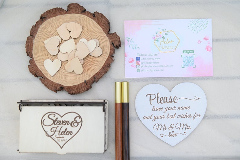 K&ouml;nnte beinhalten: Eine wei&szlig;e Holzkiste mit einem herzf&ouml;rmigen Design und dem Text "Steven & Helen September 09, 2023". Die Kiste ist umgeben von Holzh Herzen, zwei Holzstifte und einem wei&szlig;en herzf&ouml;rmigen Schild mit der Aufschrift "Bitte hinterlassen Sie Ihren Namen und Ihre besten W&uuml;nsche f&uuml;r Mr & Mrs love".