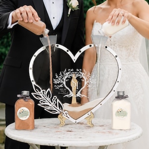 Puede incluir: Un juego de ceremonia de arena de unidad de boda en forma de corazón. El marco de acrílico transparente tiene un borde blanco y un diseño de árbol decorativo. Se están vertiendo dos botellas de arena en el marco. El juego incluye dos botellas de arena y un soporte de madera.