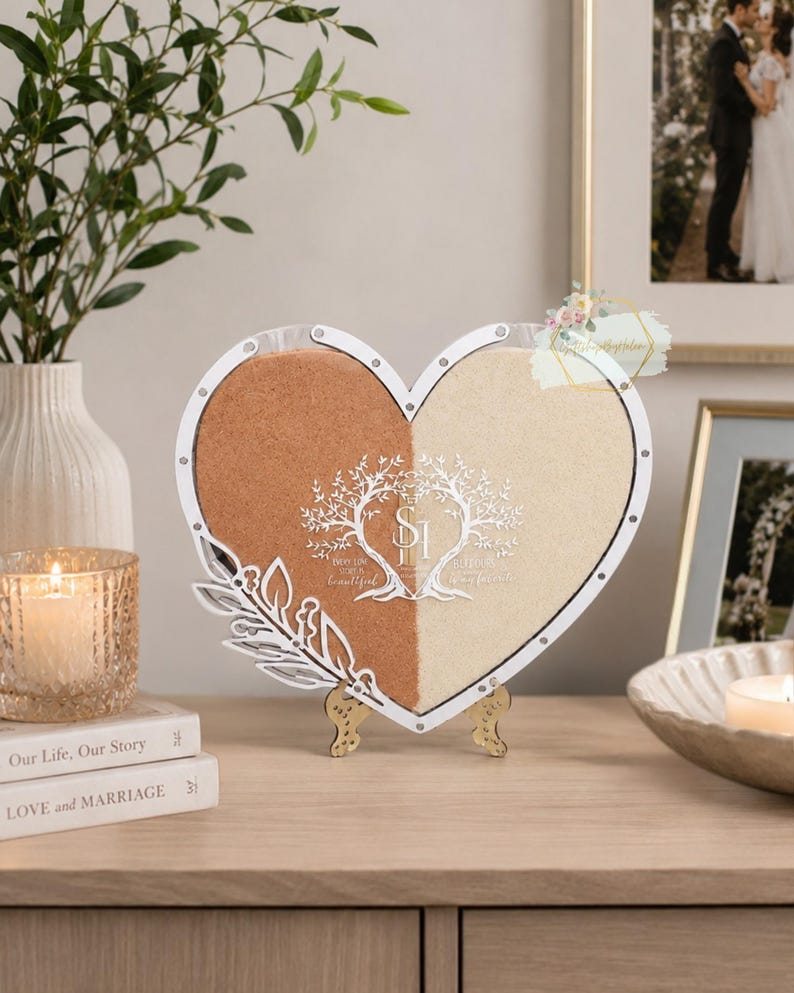 May include: A heart-shaped sand ceremony shadow box with a white frame, divided into two sections filled with tan and cream-coloured sand. The design includes a tree graphic and the text "Best Day Ever, Beautiful". It stands on a gold-coloured stand.