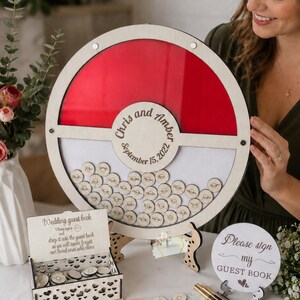 Puede incluir: Un libro de visitas de boda de madera con un dise&ntilde;o rojo y blanco. Los invitados firman peque&ntilde;os discos de madera que se dejan caer en el libro. Los nombres "Chris y Amber" y la fecha "15 de septiembre de 2022" est&aacute;n grabados. Incluye una peque&ntilde;a caja y un letrero de madera.
