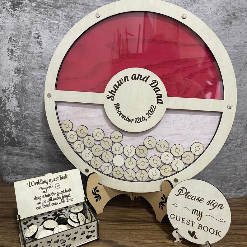 K&ouml;nnte beinhalten: Ein h&ouml;lzernes Hochzeitsg&auml;stebuch mit einem roten und wei&szlig;en Pokeball-Design. Das G&auml;stebuch hat einen Schlitz, in den G&auml;ste Holzm&auml;rkchen einwerfen k&ouml;nnen. Der Text "Shawn und Dana November 12th, 2022" ist in das G&auml;stebuch graviert.