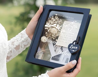 May include: A navy blue shadow box with a glass front, displaying dried flowers, a card, and the names "Steven & Helen" with the date "29.09.2025". The box is secured with a heart-shaped lock that reads "Love". The text "Happily After Ever" is visible.