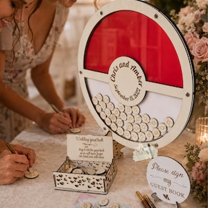 Puede incluir: Libro de visitas de boda de madera con un dise&ntilde;o circular rojo y blanco. Se muestran los nombres "Chris y Amber" y la fecha "15 de septiembre de 2022". Incluye una caja y fichas de madera para firmar.