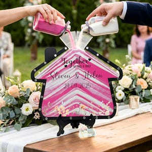 May include: A house-shaped sand ceremony frame with a black border, filled with layers of pink and white sand. The glass front features the text "Together we make a family", "Steven & Helen", and "29.09.2025" in black script, with floral accents.