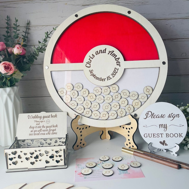 Puede incluir: Un libro de visitas de boda de madera con un dise&ntilde;o rojo y blanco. El marco circular contiene fichas de madera para que los invitados las firmen. Los nombres "Chris y Amber" y la fecha "15 de septiembre de 2022" est&aacute;n grabados. Incluye un letrero que dice "Please sign my guest book".