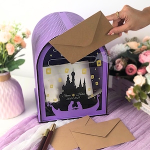 May include: A purple mailbox with a decorative design featuring a silhouette of a castle and a boat. The mailbox has a slot for inserting envelopes. The text "Steven & Helen 20.09.2025" is visible. A hand is inserting a brown envelope.