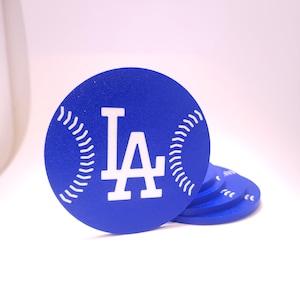 May include: Blue coasters with a baseball design. Each coaster features the white "LA" logo and white stitching details. The coasters are round and stacked, with one coaster in focus.