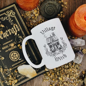 May include: White ceramic mug with a black and white illustration of a house with the text "Village Witch" on it. The mug is sitting on a wooden table with a book, candles, and other decorative items.