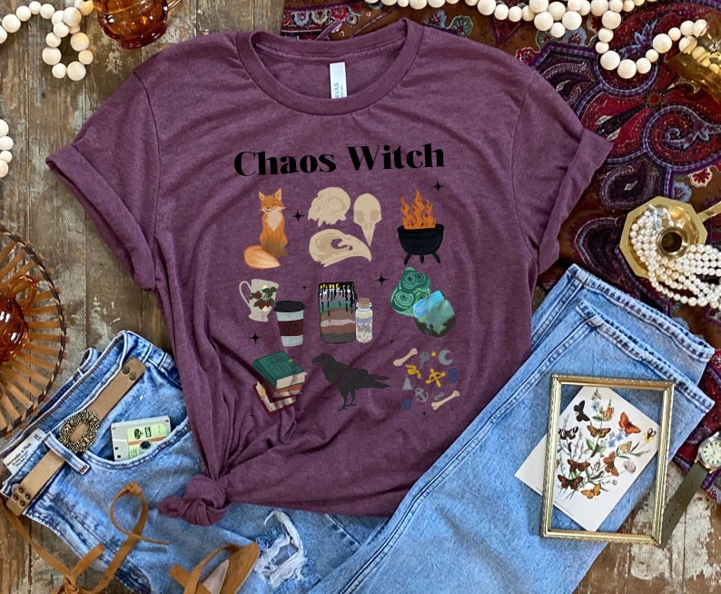 May include: A purple t-shirt with the text "Chaos Witch" printed on it. The shirt features a variety of illustrations including a fox, a cauldron, a raven, a book, and a skull.