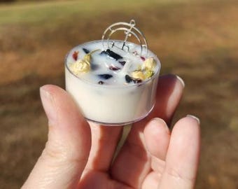 May include: A small white candle with a silver wire design on top. The candle is filled with dried flowers and black crystals.