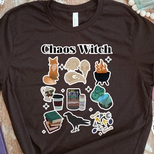 May include: Black t-shirt with the text "Chaos Witch" in white lettering. The shirt features a variety of illustrations including a fox, a skull, a cauldron, a book, a raven, and a crystal.