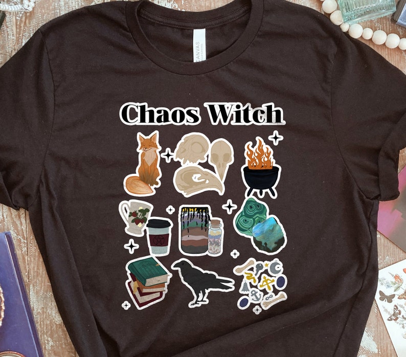 May include: Black t-shirt with the text "Chaos Witch" in white lettering. The shirt features a variety of illustrations including a fox, a skull, a cauldron, a book, a raven, and a crystal.