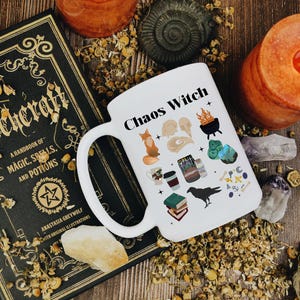 May include: A white ceramic mug with the text "Chaos Witch" and various illustrations, including a fox, a raven, and a cauldron. The mug is placed next to a book titled "Witchcraft" and a lit candle.