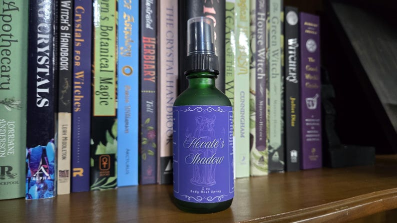 May include: A green glass bottle with a purple label that reads "Hecate's Shadow Body Mist Spray". The bottle is sitting on a wooden shelf in front of a bookshelf filled with books about witchcraft and crystals.