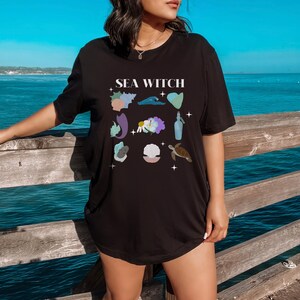 May include: Black t-shirt with the text "SEA WITCH" in white. The shirt features colorful illustrations of seashells, a mermaid tail, flowers, a bottle, and a turtle. The shirt is worn by a person on a wooden pier with the ocean in the background.