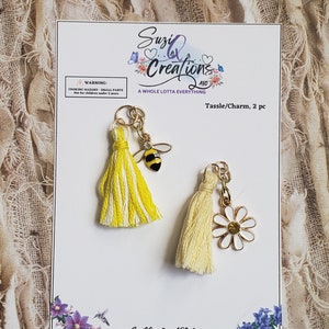 May include: Two yellow and white tassel charms with a bee and a daisy charm. The charms are attached to gold jump rings. The charms are on a white card with the text "Suzi Q Creations ABO A Whole Lotta Everything Tassle/Charm, 2 pc www.SuziQCreationsABQ.etsy.com"