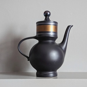 May include: A black ceramic teapot with a gold and silver band around the lid. The teapot has a round body and a spout on the side.
