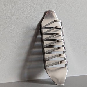 May include: A silver metal key holder with a unique design. The holder has multiple slots for keys and is shaped like a long, narrow rectangle.