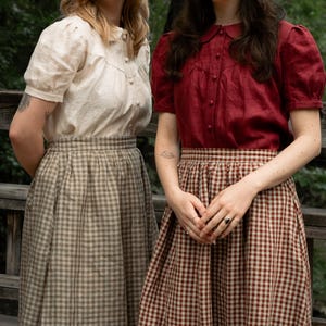 May include: Two people are seen wearing vintage-style clothing. One wears a cream blouse and a checkered skirt, the other a burgundy blouse and a checkered skirt. The outfits have a retro aesthetic, reminiscent of a bygone era.