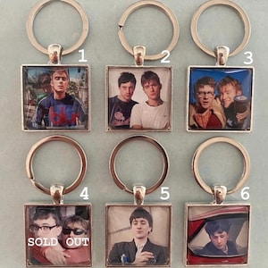May include: Six square keychains with silver-colored metal rings. Each keychain features a different image of a band member. One keychain has the words "SOLD OUT" printed on it.