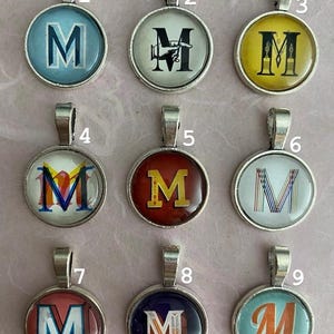 May include: Nine silver charms with different coloured glass cabochons featuring the letter M in various styles. The charms are numbered 1 through 9.