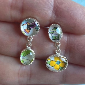 Pode incluir: Um par de brincos pendurados em tons de prata. Cada brinco apresenta um topo redondo com uma imagem colorida e um pingente menor, oval ou redondo abaixo. Um tem um beija-flor e uma borboleta, o outro um sapo e uma flor amarela.