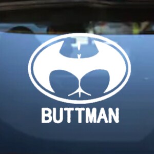 May include: White oval sticker with a humorous design. The design features a stylized bat-like shape with a buttock-like appearance, and the word "BUTTMAN" in bold, white letters below it. The sticker is affixed to a blue surface.