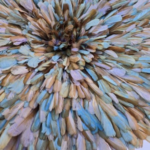 Op de afbeelding: Een close-up van een cirkelvormig decoratief stuk gemaakt van veren. De veren zijn in tinten blauw, bruin en beige, gerangschikt in een radiaal patroon. Het midden is donkerder bruin, waarbij de kleuren naar de randen toe lichter worden.