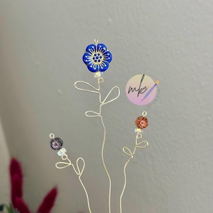 May include: Delicate floral hair accessories. The gold wire stems support blue, orange, and purple flower-shaped beads. The blue flower has a gold center. The accessories are held in a hand, with a blurred background of flowers.