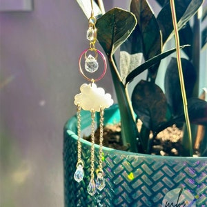 May include: Gold charm earring with a white cloud and crystal raindrops hanging from a gold chain. The earring is hanging from a plant pot.