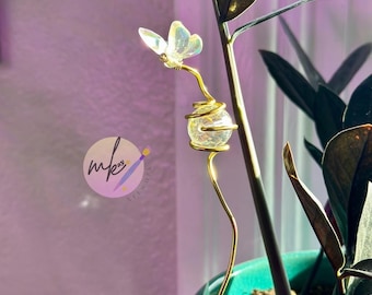 May include: A decorative plant stake featuring a butterfly design. The butterfly has iridescent wings and sits atop a gold-colored wire that wraps around a clear, spherical bead. The stake is inserted into a teal-colored pot with a green plant.
