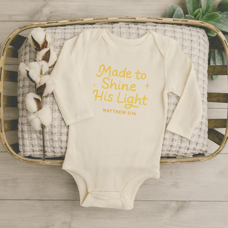 May include: Cream-colored baby onesie with long sleeves. The text "Made to Shine His Light" in yellow script is printed on the front, along with "MATTHEW 5:16". The onesie is displayed in a woven basket with a decorative cotton stem.