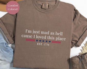 May include: A brown Comfort Colors® sweatshirt with the text "I'm just mad as hell cause I loved this place" in white. The text includes red and blue accents and the year "EST. 1776".