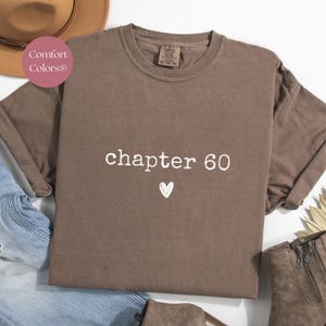 May include: A brown Comfort Colors sweatshirt with the text "chapter 60" in white, along with a small white heart. The sweatshirt is displayed with a brown hat, blue jeans, and brown boots.
