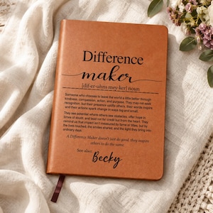 May include: A tan notebook with black text printed on the front cover. The text reads Difference maker, followed by a phonetic pronunciation, a definition of the term, and the phrase See also: Becky. A small dark brown ribbon bookmark is visible.