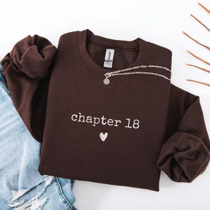 May include: A dark brown sweatshirt with the text "chapter 18" and a small heart embroidered in white. A silver necklace with a sun pendant is draped across the sweatshirt. The sweatshirt is folded on top of a pair of blue jeans.