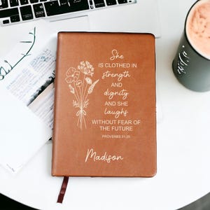 May include: A brown leather journal with the text "She is clothed in strength and dignity and she laughs without fear of the future" and "Proverbs 31:25". The journal also has the name "Madison" and a floral design.