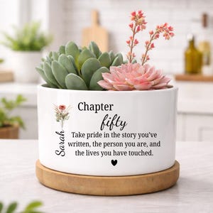 May include: White ceramic planter with a succulent arrangement. The planter has the text "Chapter fifty" and a quote, with the name "Sarah" on the side. The planter sits on a wooden base, with green and pink succulents.