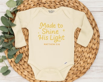 May include: Cream-colored baby onesie with long sleeves. The text "Made to Shine His Light" and "MATTHEW 5:16" are printed in gold. The onesie is displayed on a woven, circular placemat with green foliage.