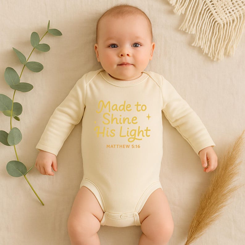 May include: A baby wearing a cream-colored onesie with long sleeves. The onesie has the text "Made to Shine His Light" in gold lettering, along with the Bible verse Matthew 5:16.