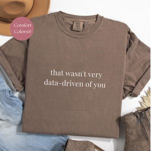 May include: A brown long-sleeve shirt with the text "that wasn't very data-driven of you" in white. The shirt is a Comfort Colors brand. A brown hat, blue jeans, and brown boots are also visible.